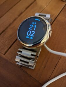Google Pixelwatch mit Metallarmband und Schutzhülle am Ladegerät hängend auf einem Holztisch. 